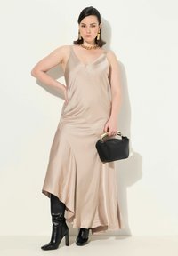Nackekleid aus Satin mit V-Ausschnitt, fließendem asymmetrischen Saum, kombiniert mit hohen schwarzen Stiefeln mit Absatz und einer kleinen schwarzen Handtasche mit goldenen Henkel.