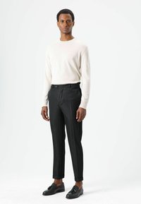 Pull en maille beige clair, pantalons noirs sur mesure avec une finition polie, et chaussures en cuir noir. Modèle debout sur un fond blanc.