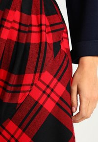 Punane ja must ruudulise mustriga seelik, millel on pleeditud disain, valmistatud tekstuuriga kangast. Käsi on paigutatud talje lähedale.