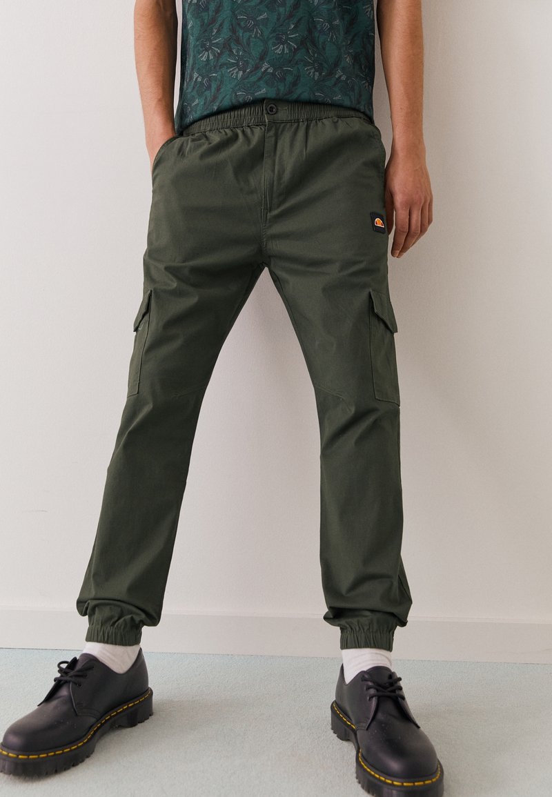 Una persona está de pie con las manos en los bolsillos, vistiendo pantalones cargo oliva y una camisa verde de estampado, combinada con zapatos negros y calcetines blancos.