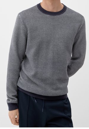 Mand iført en grå tekstureret langærmet sweater med mørke manchetter og krave, kombineret med mørkeblå bukser mod en ensfarvet baggrund.