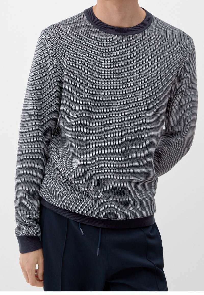 Homme portant un pull gris texturé à manches longues avec des poignets et un col foncés, associé à un pantalon bleu foncé sur un fond uni.