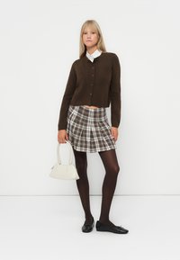 Γυναίκα που φοράει καφετί ζακέτα, λευκό πουκάμισο με γιακά, καρό φούστα, μαύρες καλσόν, μαύρα φλατ παπούτσια, κρατώντας μια λευκή τσάντα, στέκεται μπροστά σε λευκό φόντο.