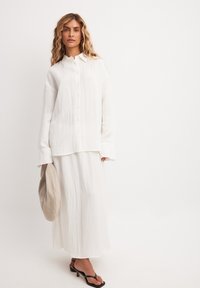 Witte overhemd met lange mouwen, gecombineerd met een lange witte rok. Zwarte strappy sandaaltjes en een beige tas maken de outfit compleet.