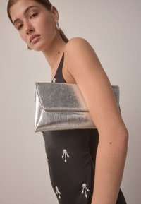 Ezüst fémes kuplung texturált felülettel, hajtogatott fedéllel, fekete ruhához tartva, melyet gyöngy díszítések ékesítenek.