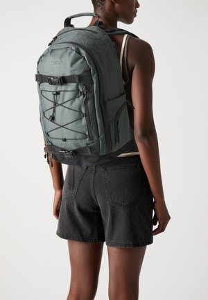 Mochila de nylon verde con múltiples compartimentos, correas ajustables y un diseño de cordón elástico en la parte frontal. Textura suave y acentos de hardware mínimos.