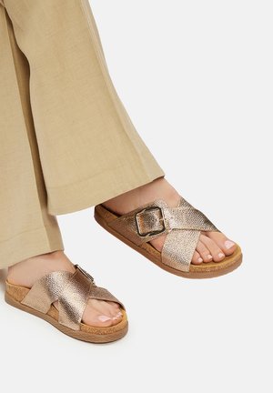 Sandalias planas con tiras y acabado metálico texturizado en color oro rosa, con una gran hebilla cuadrada y suela de corcho marrón claro.