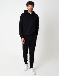 Schwarzer Hoodie mit Kängurutasche vorne, Kordelzügen und kleinem Logo, kombiniert mit passenden Jogginghosen und weißen Turnschuhen.