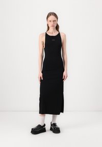 Abito halter nero a coste con una fessura laterale, realizzato in tessuto morbido. Presenta un piccolo logo sul petto ed è abbinato a scarpe nere spesse.
