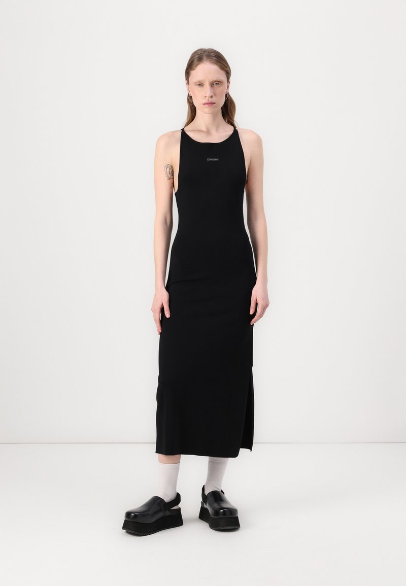 Abito halter nero a coste con una fessura laterale, realizzato in tessuto morbido. Presenta un piccolo logo sul petto ed è abbinato a scarpe nere spesse.