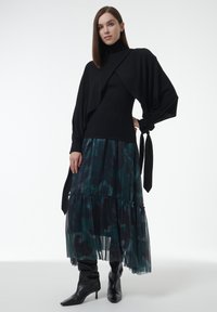 Zwart hemd met lange mouwen en gedrapeerd ontwerp, gecombineerd met een geplooide groene rok met bloemendessin en kniehoge zwarte laarzen.