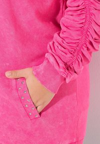 Sudadera rosa con detalles de mangas fruncidas, tejido texturizado, puños acanalados y un bolsillo adornado con acentos plateados en forma de estrella.