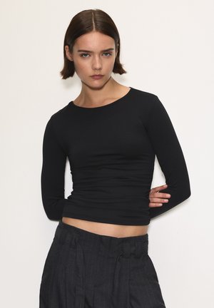 Tânără cu părul scurt și brunet, purtând un top negru mulat cu mânecă lungă și pantaloni în dungi întunecate cu talie înaltă, stând în fața unui fundal alb.