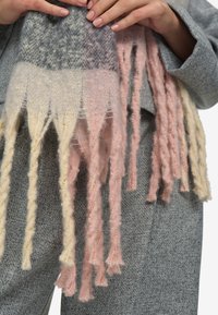 Mani che tengono una sciarpa in lana morbida e frangiata con blocchi rosa, grigi e crema sopra pantaloni grigi texture.