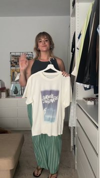 Camiseta crema con un gráfico que dice "Seashells Juneports", colgando de una percha blanca. Presenta mangas cortas y un cuello redondo.