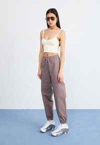 Mlada žena stoji samouvjereno u svijetloj majici bez rukava i širokim sivim hlačama, noseći sunčane naočale i stylish tenisice na plavoj pozadini.
