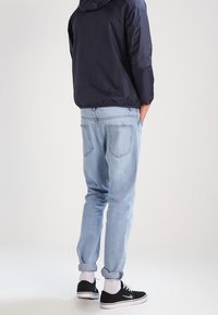 Giacca con cappuccio navy, jeans tapered azzurri chiari, sneakers nere con suola bianca e orli arrotolati. Sfondo neutro, focus sui dettagli dell'abbigliamento.