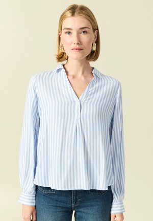 Donna con capelli biondi corti che indossa orecchini dorati, una blusa a righe verticali azzurre e bianche e jeans blu scuro su sfondo beige.