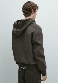 Bruine capuchontrui met een gestructureerde afwerking; ontspannen pasvorm, geribbelde manchetten en zoom; gedragen met meerdere lagen kleding eronder.