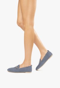 Blaue Wildlederslipper mit flacher Sohle, ausgestattet mit einem dekorativen Metallelement und einem offenen Rückendesign für ein einfaches An- und Ausziehen.