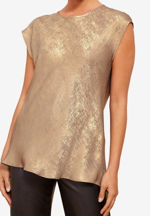 Frau trägt eine ärmellose, metallisch goldene, strukturierte Bluse kombiniert mit schwarzen Hosen, fotografiert von Hals bis Hüfte.