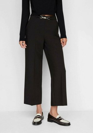 Femme portant un pantalon noir court à jambes larges avec une ceinture à boucle argentée, associé à des mocassins noirs et blancs et un haut noir à manches longues.