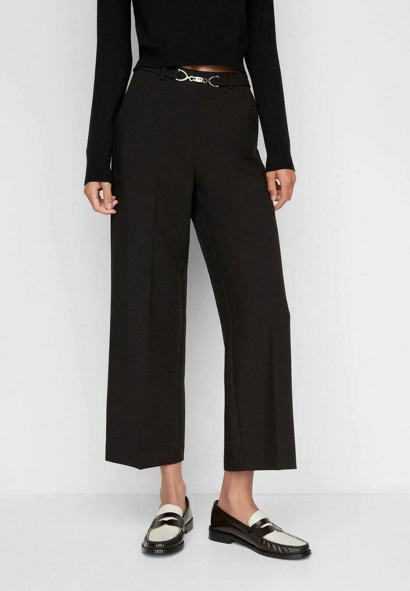Femme portant un pantalon noir court à jambes larges avec une ceinture à boucle argentée, associé à des mocassins noirs et blancs et un haut noir à manches longues.