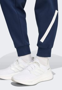 Navy athletic pants with white side stripe paired with white running shoes. Shoes feature textured fabric and three stripe branding.