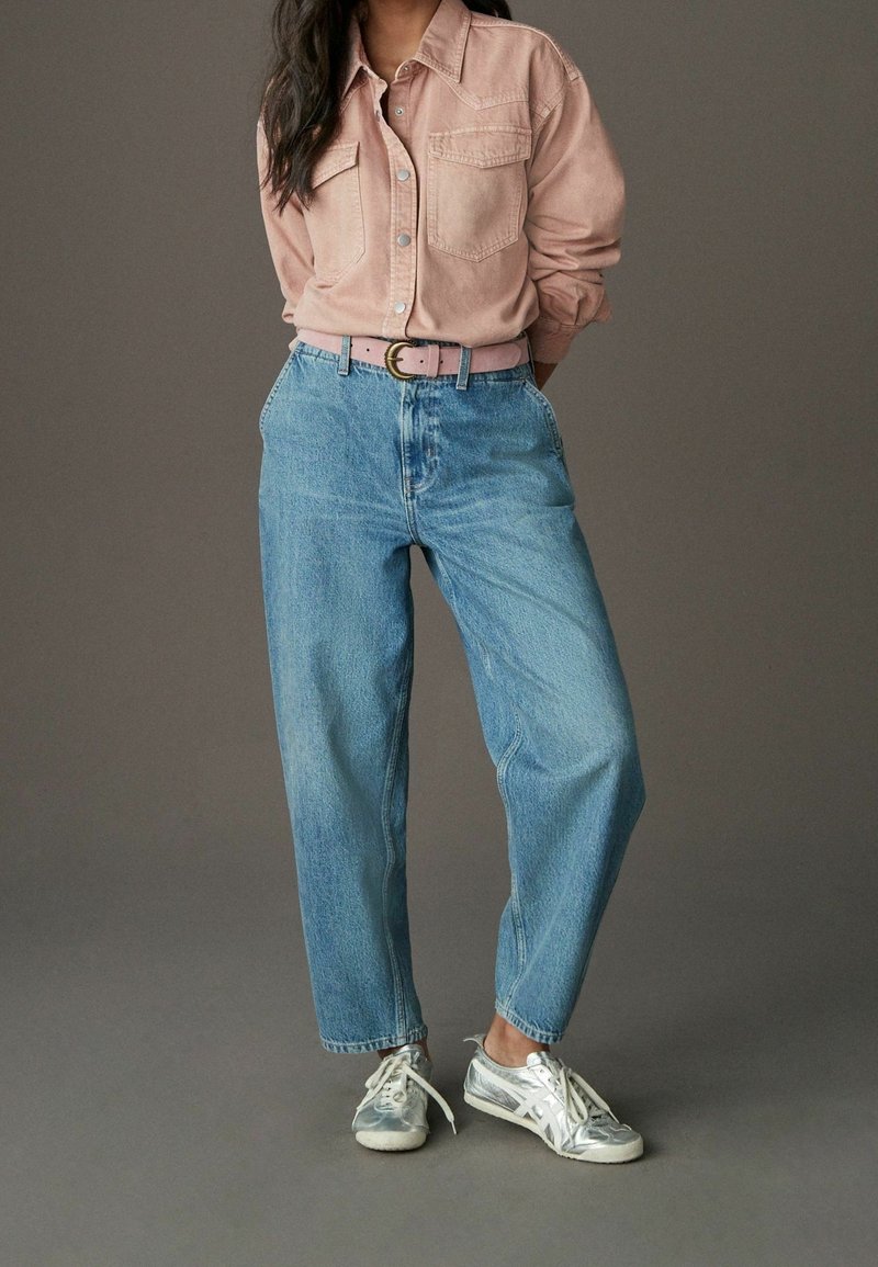 Personne portant une chemise à boutons rose clair, un jean taille haute bleu avec ceinture rose, et des baskets argentées, debout sur un sol gris.