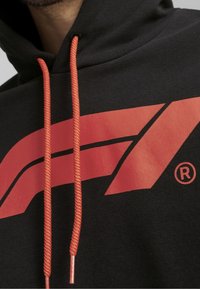 Felpa nera con cappuccio caratterizzata da un logo rosso oversize, coulisse arancione con estremità in metallo e una texture morbida. La vista ravvicinata mette in evidenza i dettagli del design.