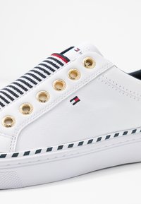 Baskets blanches avec un panneau en tissu rayé, œillets dorés, et une semelle à rayures noires et blanches. Matière en cuir texturé.