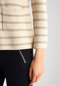 Maglione lavorato a maglia di colore crema con righe orizzontali marroni, dotato di una tasca frontale e polsini a costine, abbinato a pantaloni scuri con zip dorata.