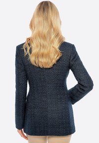 Dunkelblazer aus strukturiertem Stoff mit einem Karomuster. Hat lange Ärmel und eine taillierte Silhouette, ohne sichtbare Hardware.