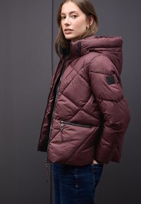 Bordeauxfarbene, gesteppte Pufferjacke mit Kapuze, diagonal gesteppt, schwarzen Reißverschlusstaschen und einem markierten Patch am Ärmel.