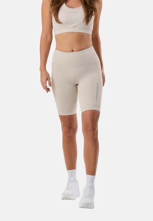 Donna che indossa pantaloncini da biker beige a vita alta, reggiseno sportivo abbinato, calzini bianchi e scarpe da ginnastica bianche in piedi davanti a uno sfondo semplice.