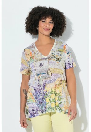 Kvinde med krøllet hår iført en farverig T-shirt med blomster- og sommerfuglemønster med V-hals og lysegule bukser, smilende blidt.
