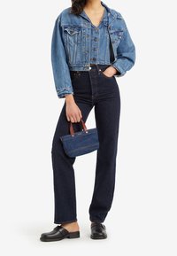 Gekürzte blaue Jeansjacke mit Druckknopfverschluss, kombiniert mit dunkelblauen, hochgeschnittenen Jeans. Das Model hält eine kleine Jeanstasche mit Ledergriffen.