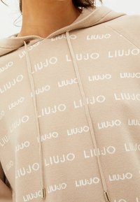 Beige hoodie met een all-over witte "LIU JO" logo print, voorzien van een capuchon met trekkoord en een grofgebreide stof. Details in goudkleurige hardware.