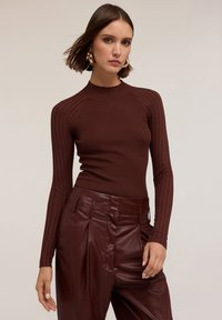 Donna che indossa un top marrone a maniche lunghe a costine e pantaloni in pelle nera a vita alta, con capelli corti e grandi orecchini a cerchio dorati.