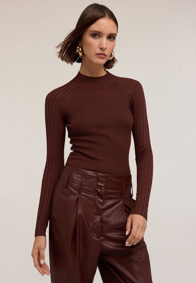 Donna che indossa un top marrone a maniche lunghe a costine e pantaloni in pelle nera a vita alta, con capelli corti e grandi orecchini a cerchio dorati.