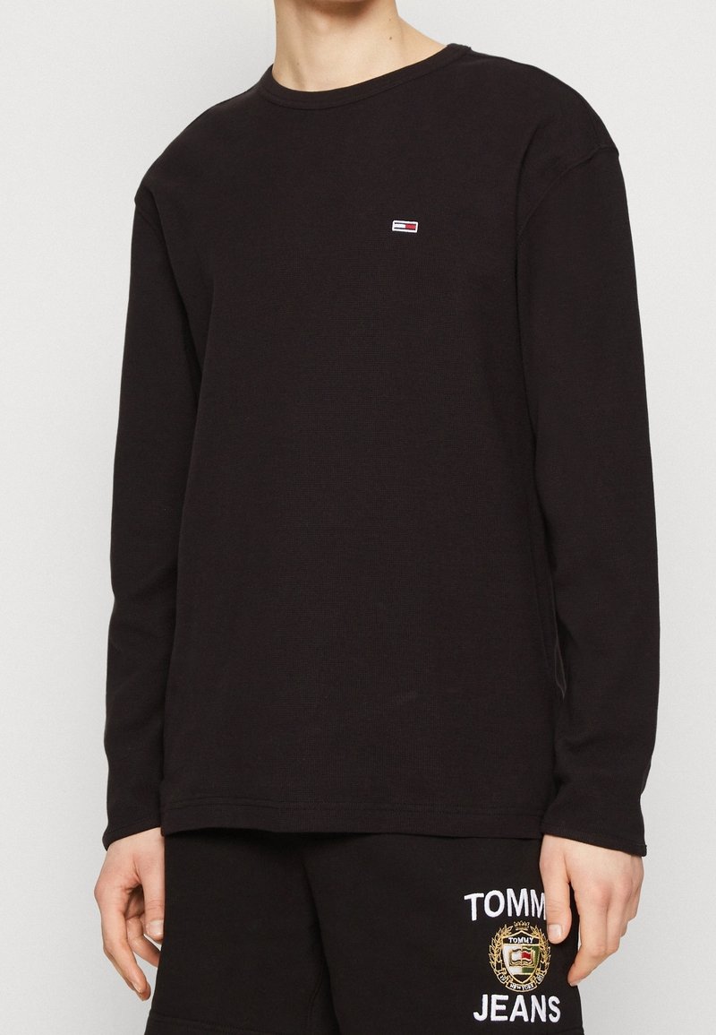 Svart långärmad t-shirt i mjukt stickat material, med en liten röd och vit logotyp på bröstet. Rund halsringning och avslappnad passform.