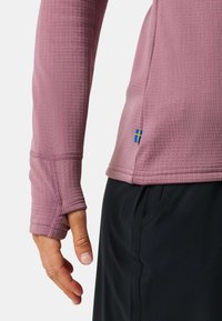 Long-sleeve athletic top in muted pink, featuring a textured fabric, thumb holes, and a small Swedish flag accent near the hem. Paired with black shorts.