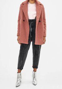 Cappotto di grandi dimensioni, rosa e texture con rever e doppi bottoni indossato sopra un top rosa chiaro e pantaloni di denim neri a vita alta. Stivali alla caviglia argento.