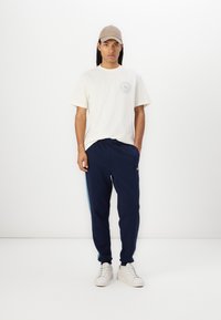 Tricou crem deschis cu un logo circular, pantaloni bleumarin cu dungi laterale în nuanța bleu deschis și adidași albi, toate într-un stil minimal.