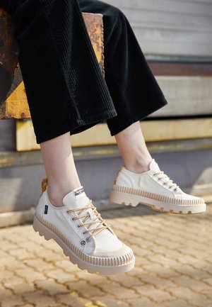 Osoba nosząca białe sneakersy na grubej podeszwie z beżowymi sznurówkami i czarne szerokie spodnie, siedząca na zewnątrz na utwardzonym podłożu z lekko uniesionymi stopami.