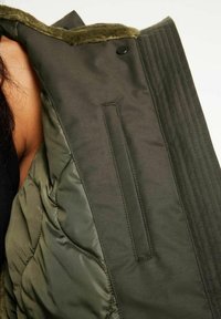 Grön vadderad jacka med en mjuk pälskrage, slät tyg och en smal horisontell ribbad struktur. Inkluderar en ficka med dragkedja.