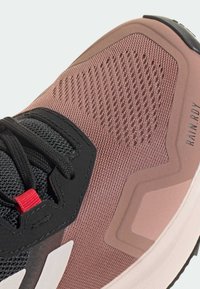 Buty sportowe z różową siateczką, czarnymi sznurówkami i czerwonym akcentem; posiadają teksturowane elementy wzornicze oraz elastyczną gumową podeszwę.