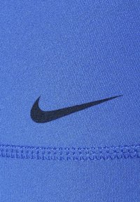Modrá sportovní tkanina s hladkým povrchem, na které je černé logo Nike swoosh. Obsahuje prošívaný šev podél okraje.