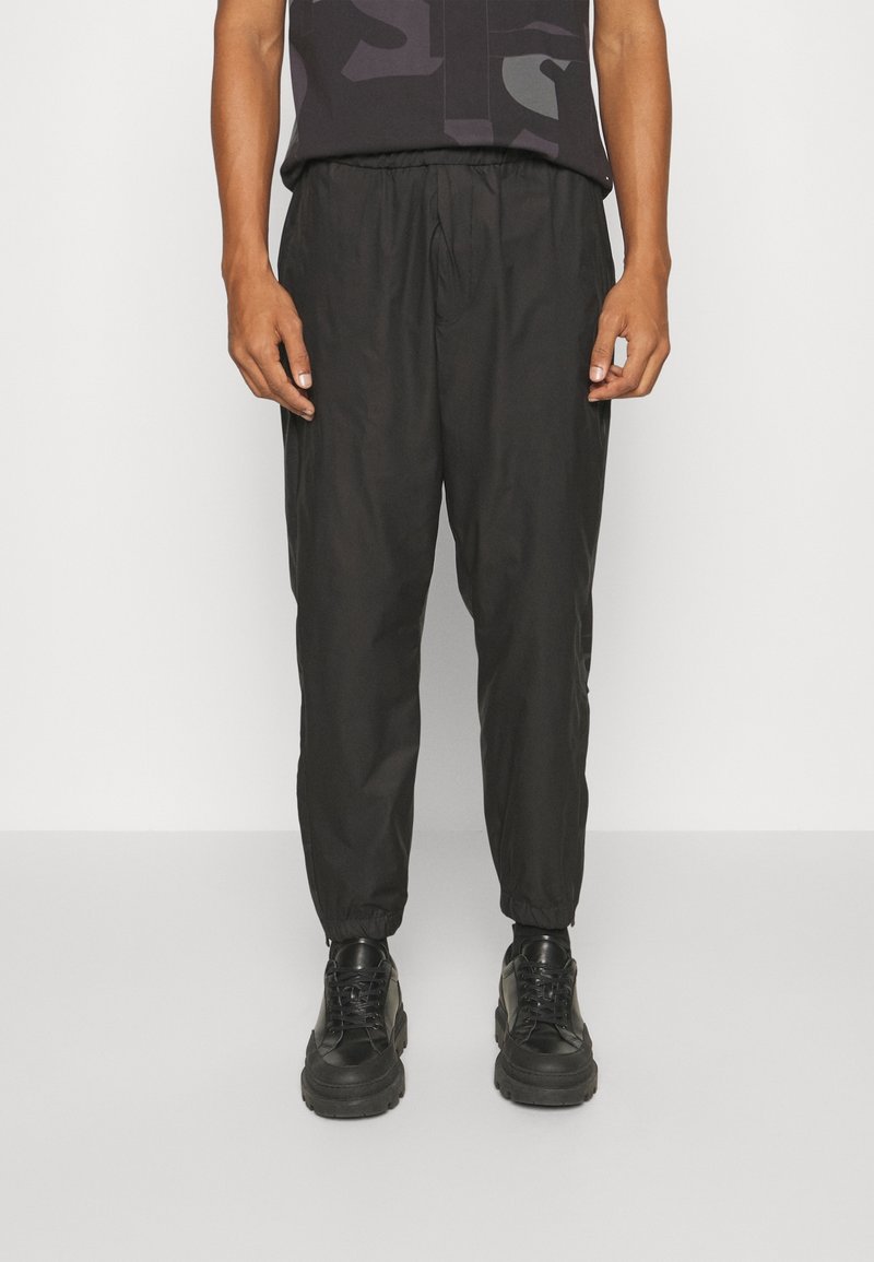 Černé tapered tepláky s elastickým pasem, hladkým povrchem a žebrovanými manžetami, doplněné černými šněrovacími teniskami.