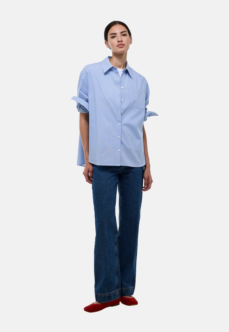 Chemise oversize rayée bleue et blanche avec un col, boutonnière à l'avant, manches retroussées ; portée avec un jean boyfriend bleu évasé et des chaussures rouges.
